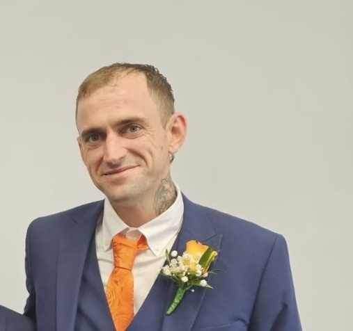 A photograph of a man wearing a navy suit with an orange tie. 